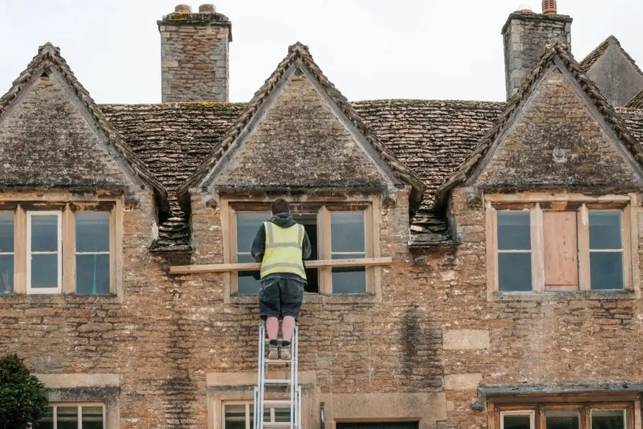 A construction worker on a ladder repairs a window on a stone building.